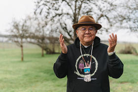 Indigenous man wearing black hoodie, hat and necklace with hands raised